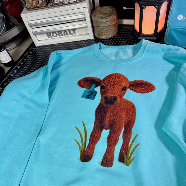 Light blue t-shirt with a faux knitted brown brangus calf design on a table.