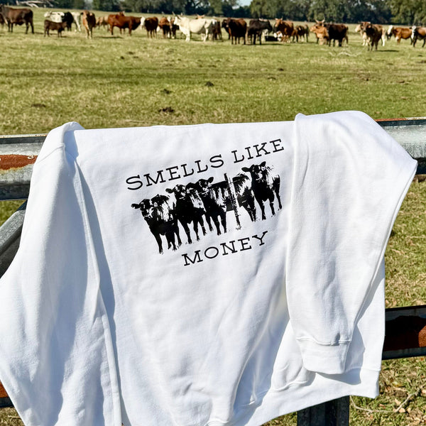 White sweatshirt with 'Smells Like Money' and cow graphic on a fence in a field with cows.