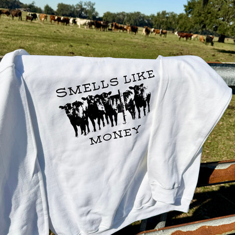 White sweatshirt with 'Smells Like Money' graphic on a wooden bench with cows in the background.