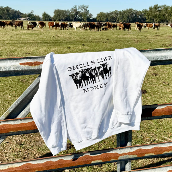 White sweatshirt with 'SMELLS LIKE MONEY' text and cow graphic hanging on a fence with cows in the background.