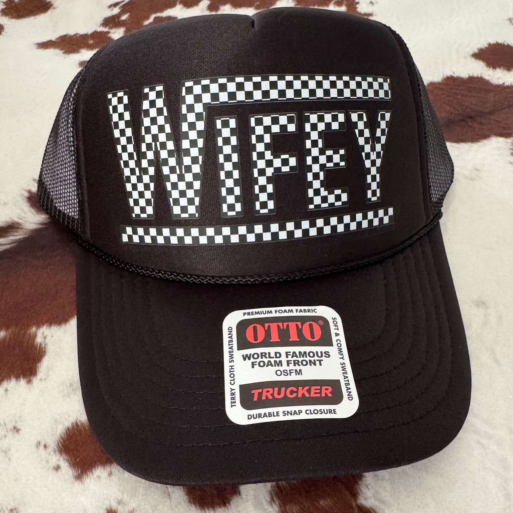 Black trucker cap with 'Wifey' text on a checkered pattern, brand 'OTTO' visible, on a textured brown and white background.