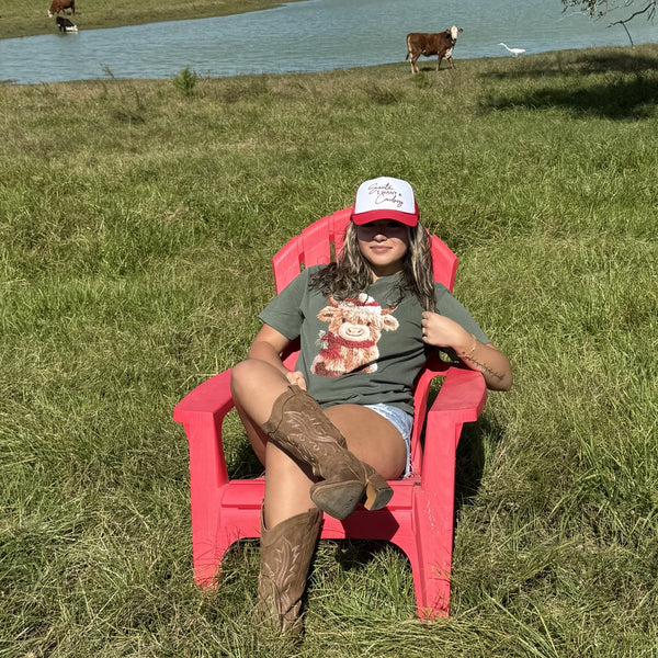 Person wearing Christmas Cow graphic tee & cowgirl trucker hat sitting on a red chair in a grassy field with cows in the background