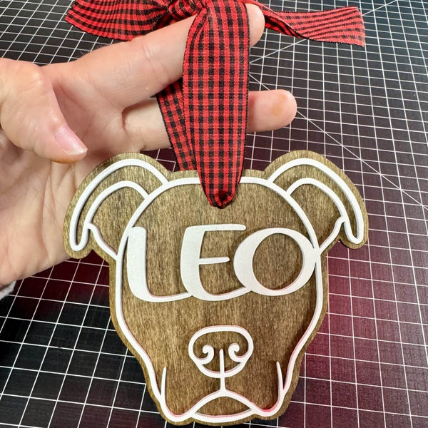 Wooden American Pitbull dog-shaped ornament with 'LEO' on it, held by a hand with a red checkered ribbon.