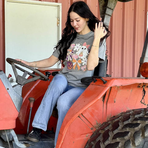 Woman sitting on an orange tractor with a  gray coquette style ghost graphic t-shirt.