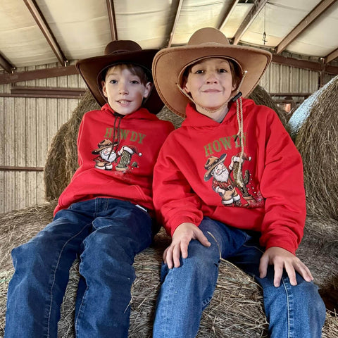 Two children wearing red hoodies with cowboy santa graphics sitting on hay bales.