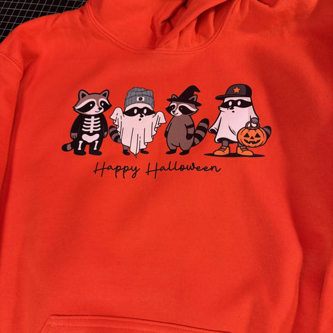 Orange youth hoodie with four raccoons in Halloween costumes and text “Happy Halloween.”