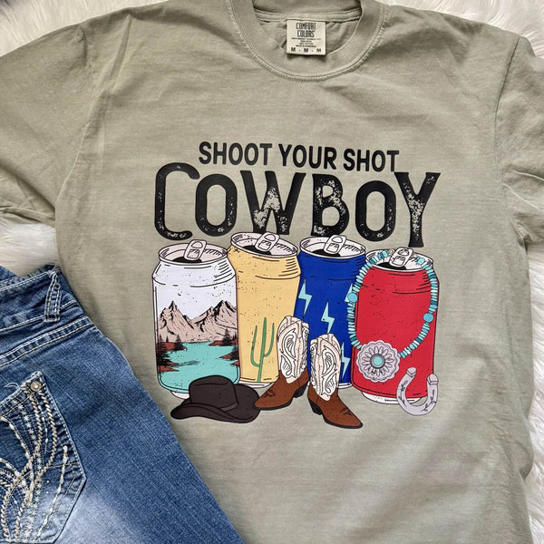 Khaki color t-shirt with 'Shoot Your Shot Cowboy' graphic featuring cans and boots, paired with jeans.