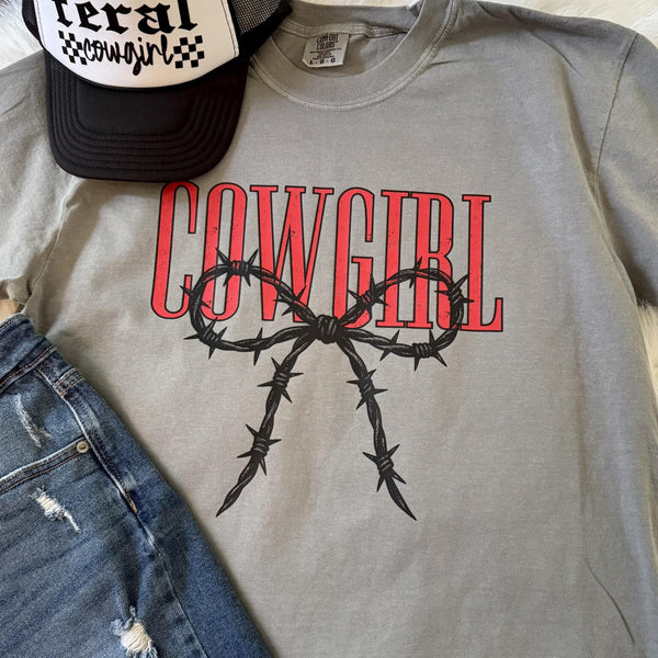 Comfort Colors tee with the word “Cowgirl” in distressed red letters and a black barbed-wire bow graphic, styled with denim and a trucker hat.