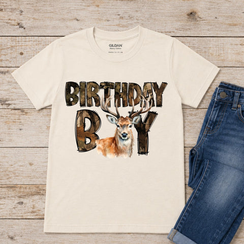 An off-white Gildan baby cotton t-shirt with a 'BIRTHDAY BOY' graphic featuring a deer, styled in a flat lay on a rustic wood background next to a pair of blue jeans.