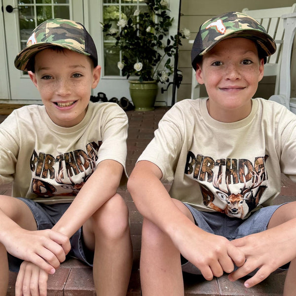 Two boys sitting side-by-side on a porch, wearing matching beige graphic t-shirts with a deer design and 'BIRTHDAY BOY' text, camouflage baseball caps, and denim shorts.