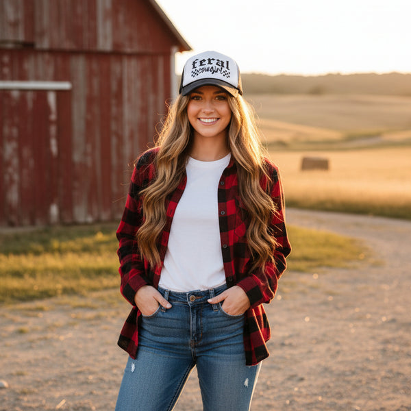 Woman wearing a cowgirl trucker hat and jeans standing in front of a red barn.