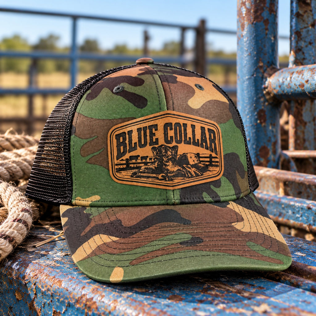 Blue Collar camo trucker hat with leather patch displayed on a rustic cattle ranch gate