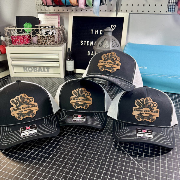 Black and white Richardson 112 trucker hats with brown leather mechanic tool patches, featuring wrenches, gears and automotive tools in a rustic workshop setting