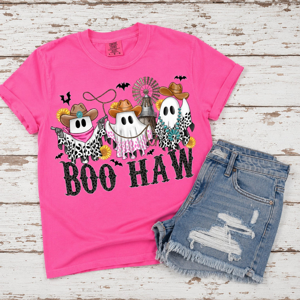 A bright pink graphic T-shirt laid flat on a whitewashed wood background, paired with light blue distressed denim shorts. The shirt features a Halloween-themed western ghost design with cowboy hats, leopard-print details, and the text 'BOO HAW'.