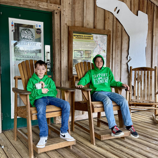 Two children in green farm hoodies sitting on wooden chairs on a wooden porch.