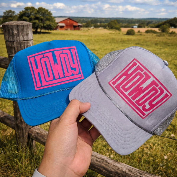 Blue and gray Howdy trucker hats with pink graphic held in front of a rustic country barn landscape