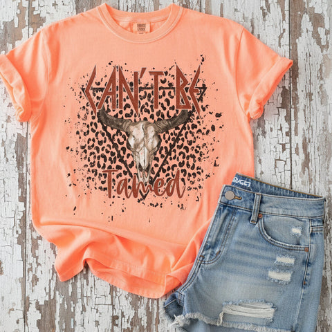 Neon Cantaloupe Comfort Colors brand t-shirt with a "Can't be Tamed" cow skull graphic design on a white wooden background