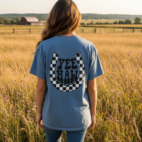 Woman wearing blue t-shirt with checkered 'Yee Haw' horseshoe design standing in a country field