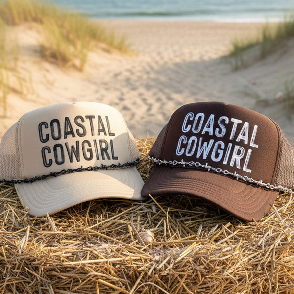 Two 'Coastal Cowgirl' hats on a beach with sand and grass in the background.