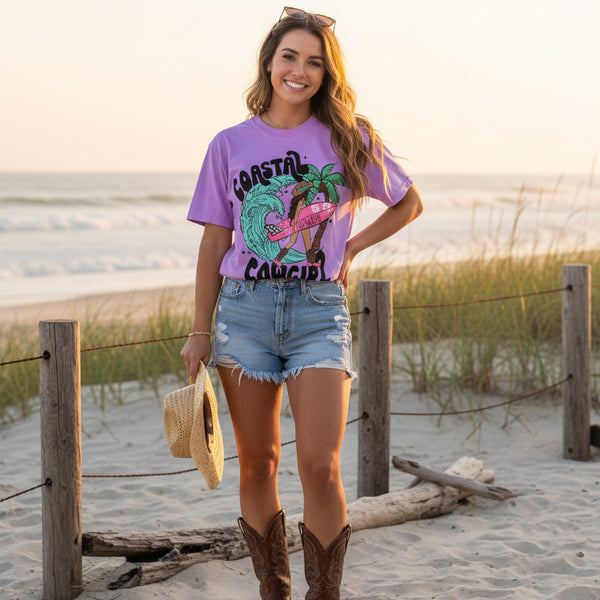 Coastal cowgirl graphic tee in neon violet worn on beach with denim shorts and cowboy boots, southern country summer outfit
