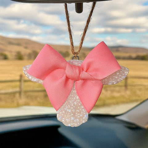 Scented cow wearing a pink bow with white beads hanging from a car's rearview mirror with a scenic background.
