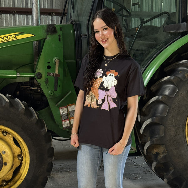 Person wearing a black t-shirt with coquette cowgirl ghost characters in front of a green tractor.