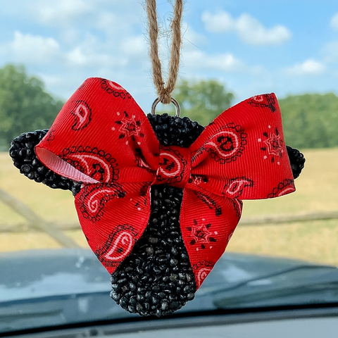 Decorative red and black paisley bow air freshener hanging on a car's rearview mirror with a blurred outdoor background.