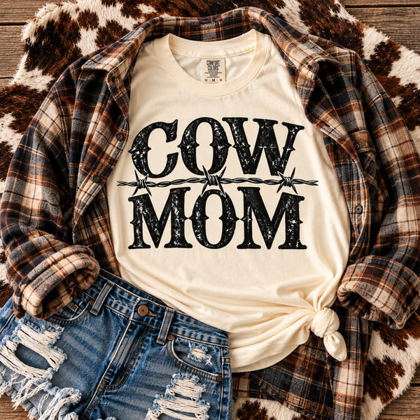 Ivory Cow Mom western graphic t-shirt layered with brown plaid flannel and distressed denim shorts on rustic cowhide background.