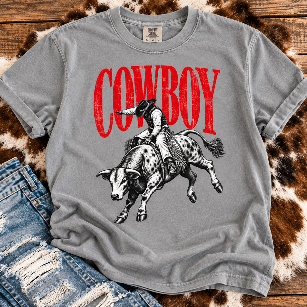 Gray Comfort Colors retro rodeo graphic tee with cowboy riding a bucking bull, styled with distressed denim, cowhide, and rustic wood background.