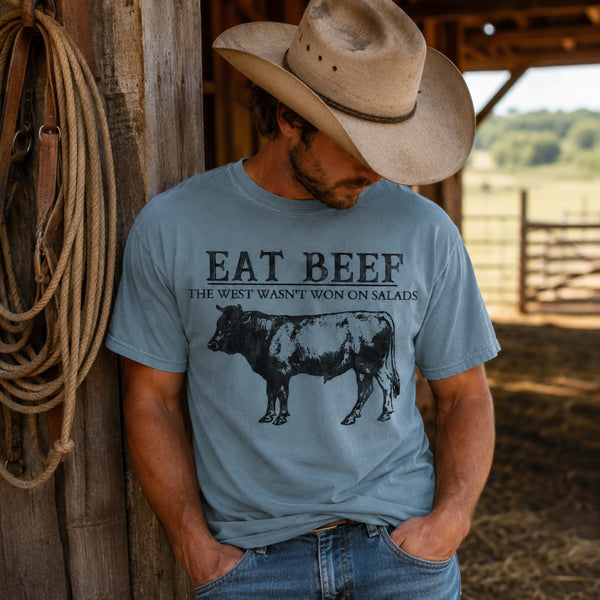 A man wearing a light blue graphic t-shirt and a cowboy hat, leaning against a rustic wooden barn post with a coiled rope. The t-shirt features a black graphic of a cow and the text 'EAT BEEF THE WEST WASN'T WON ON SALADS'.