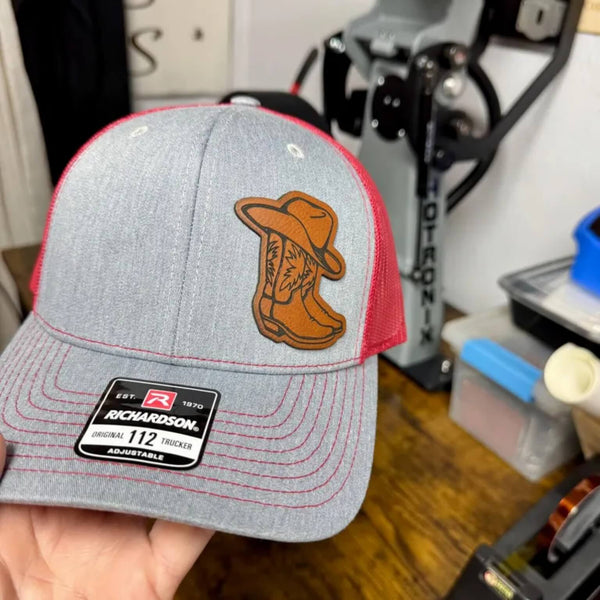 Gray and red trucker hat with a leather boot patch held in front of a workshop background.