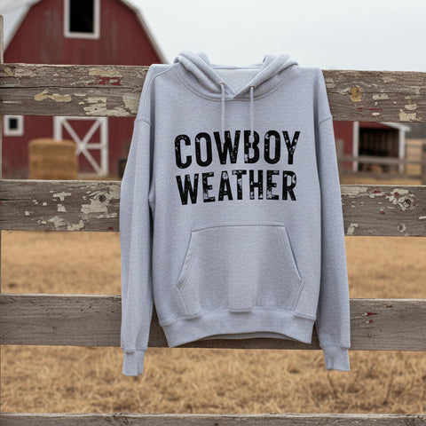 Gray hoodie with 'Cowboy Weather' text hanging on a barnyard fence