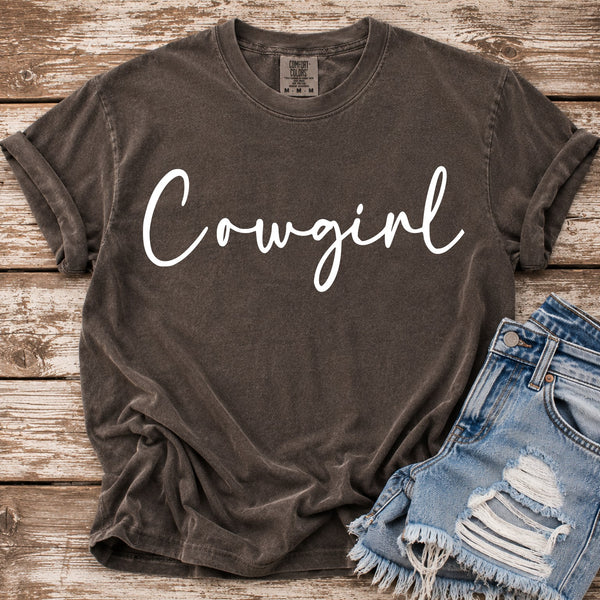 A vintage washed brown Comfort Colors t-shirt with 'Cowgirl' printed in white script across the chest, styled flat on a rustic wooden background next to a pair of distressed denim shorts.