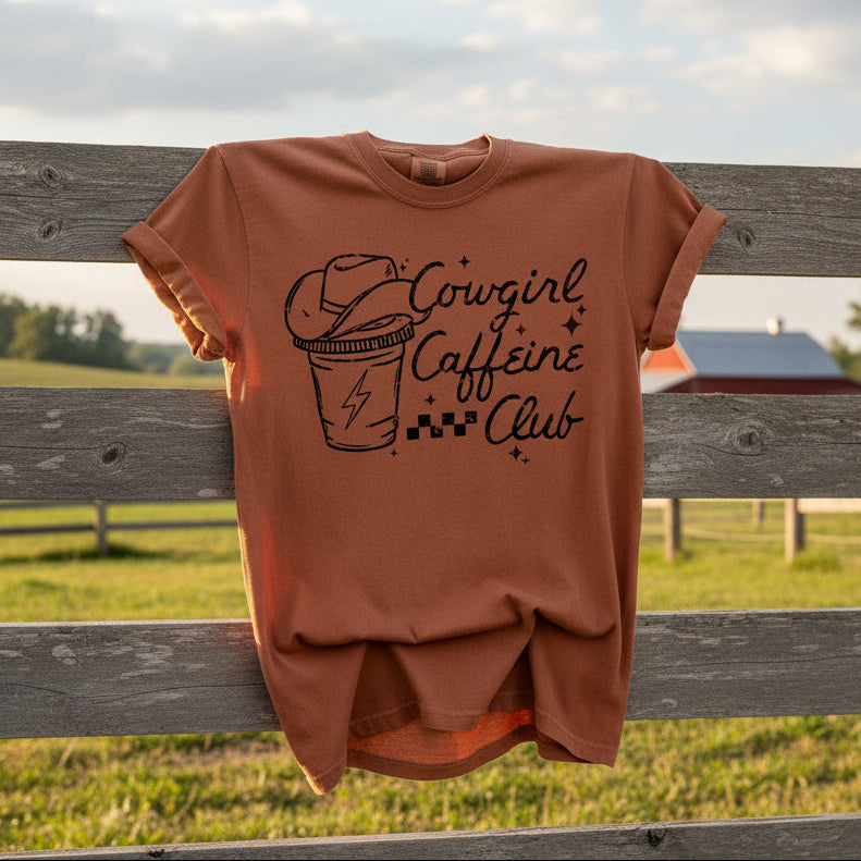 Brown t-shirt with 'Cowgirl Caffeine Club' text and graphic hanging on barnyard fence
