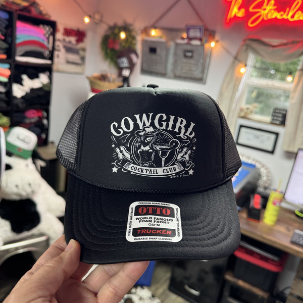Black trucker hat with 'Cowgirl Cocktail Club' design held in a store setting.