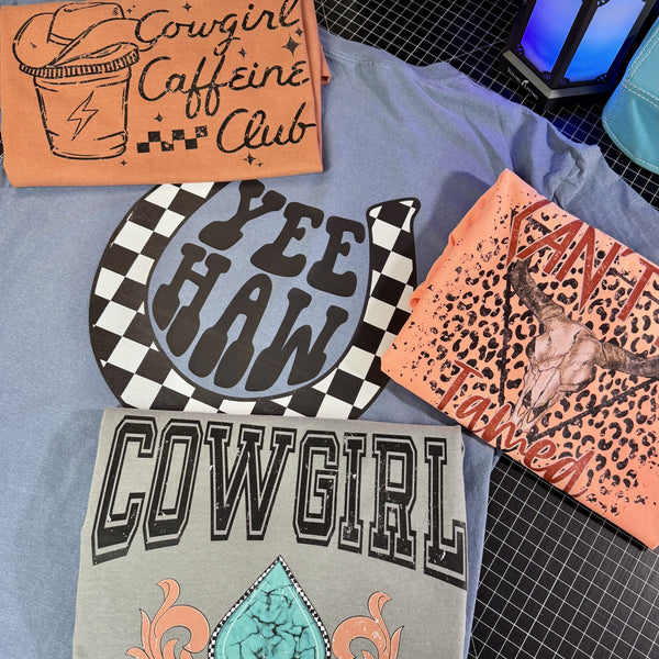 Collection of graphic tee shirts with cowgirl-themed designs on a checkered surface.