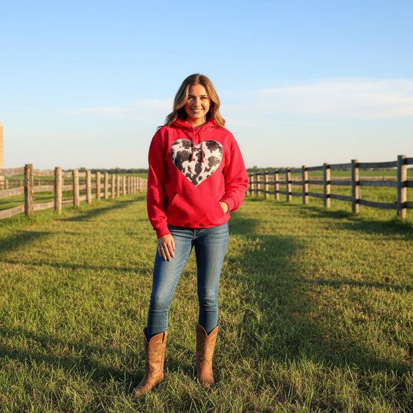 Woman wearing a red hoodie with a heart design in a grassy field with wooden fences and a barn in the background.