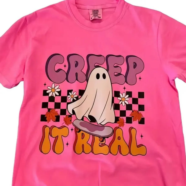 A bright pink short-sleeve t-shirt featuring a retro-style graphic with the text 'CREEP IT REAL' in purple and orange bubble letters, a cartoon ghost on a skateboard, and small white flowers over a black-and-white checkerboard pattern.