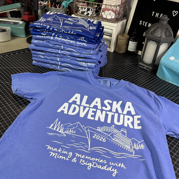 Blue t-shirt with 'Alaska Adventure 2026' design on a table with stacks of similar shirts.