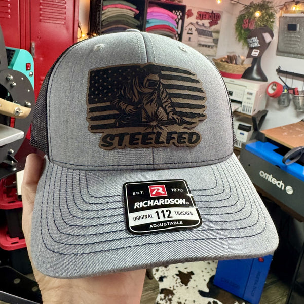 Gray Richardson 112 trucker hat with custom black leatherette patch featuring a welder in front of an American flag and surname text — handmade welding hat, patriotic welder cap, blue collar workwear gift, laser engraved patch hat, industrial tradesman apparel