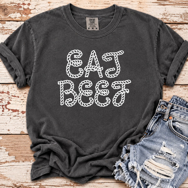 A dark charcoal gray t-shirt with 'EAT BEEF' printed in white rope-style lettering, styled flat on a distressed white and brown wood background next to a pair of distressed denim shorts.