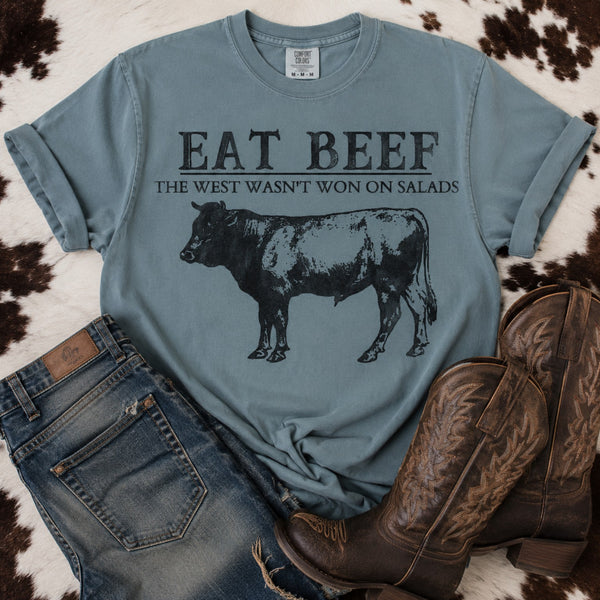 Vintage Eat Beef graphic printed on a Comfort Colors Ice Blue unisex tee with a black cow illustration and "The West Wasn't Won on Salads" slogan.