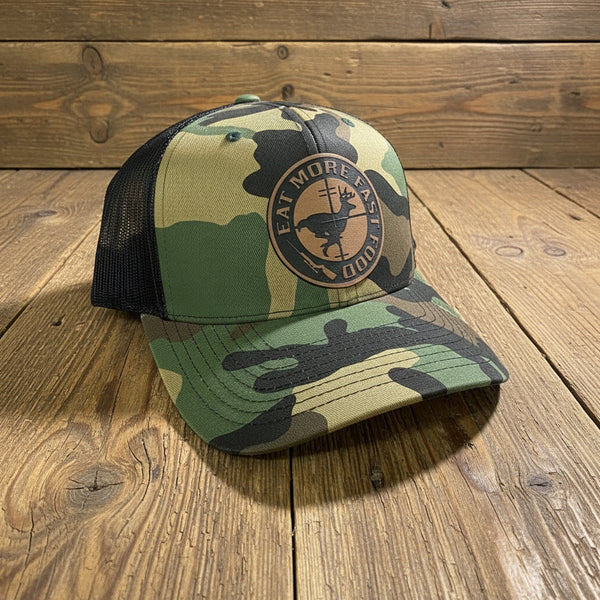 Eat More Fast Food deer hunter camo trucker hat with engraved buck patch, rustic outdoorsman hunting cap on wooden background.