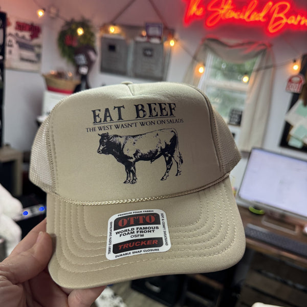 Beige cap with 'Eat Beef' text and cow graphic held in front of a casual indoor setting.