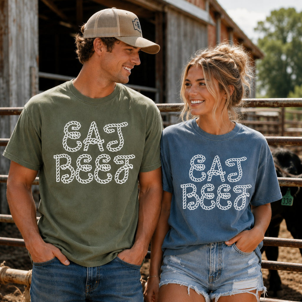 A man and a woman standing outdoors in front of a rustic barn and fence, wearing matching graphic T-shirts that read 'EAT BEEF' in a white rope-style font. The man wears an olive green shirt, and the woman wears a dusty blue shirt.