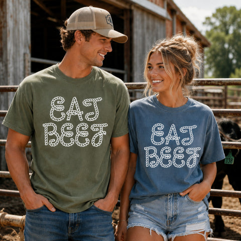 A man and a woman standing outdoors in front of a rustic barn and fence, wearing matching graphic T-shirts that read 'EAT BEEF' in a white rope-style font. The man wears an olive green shirt, and the woman wears a dusty blue shirt.