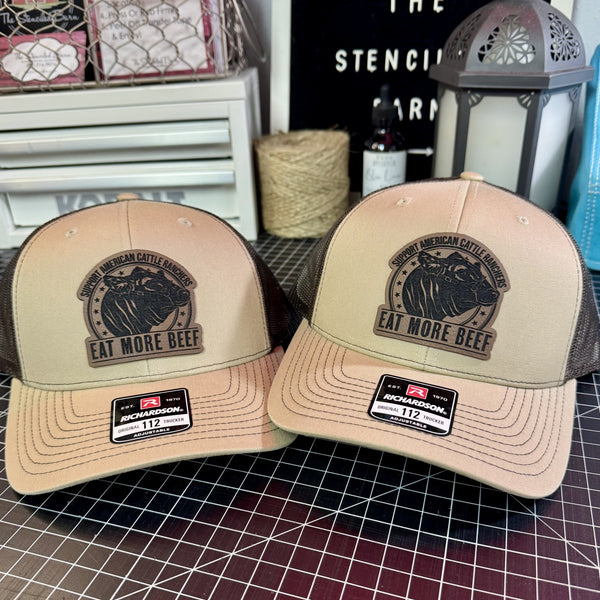 Two beige trucker hats with 'Eat More Beef' logo on a grid surface.