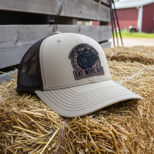 Beige trucker hat with a Eat More Beef Cattle Rancher logo sitting on a haystack in a barnyard