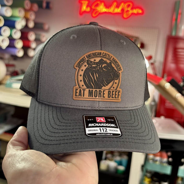 Gray trucker hat with a Eat More Beef logo and text held in front of a colorful background.
