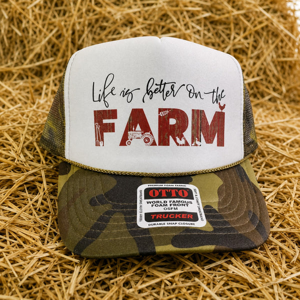 Farm life camo trucker hat with white foam front, distressed red farm graphic and rope detail displayed on hay